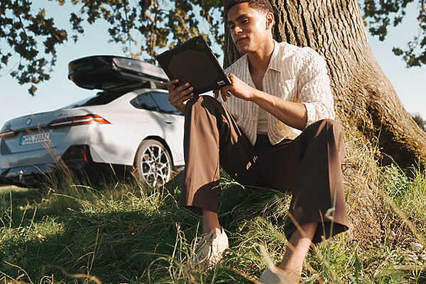 My BMW App – unterwegs verbunden Frau mit Tablet an einem Baum neben einem geparkten BMW mit Dachbox.
