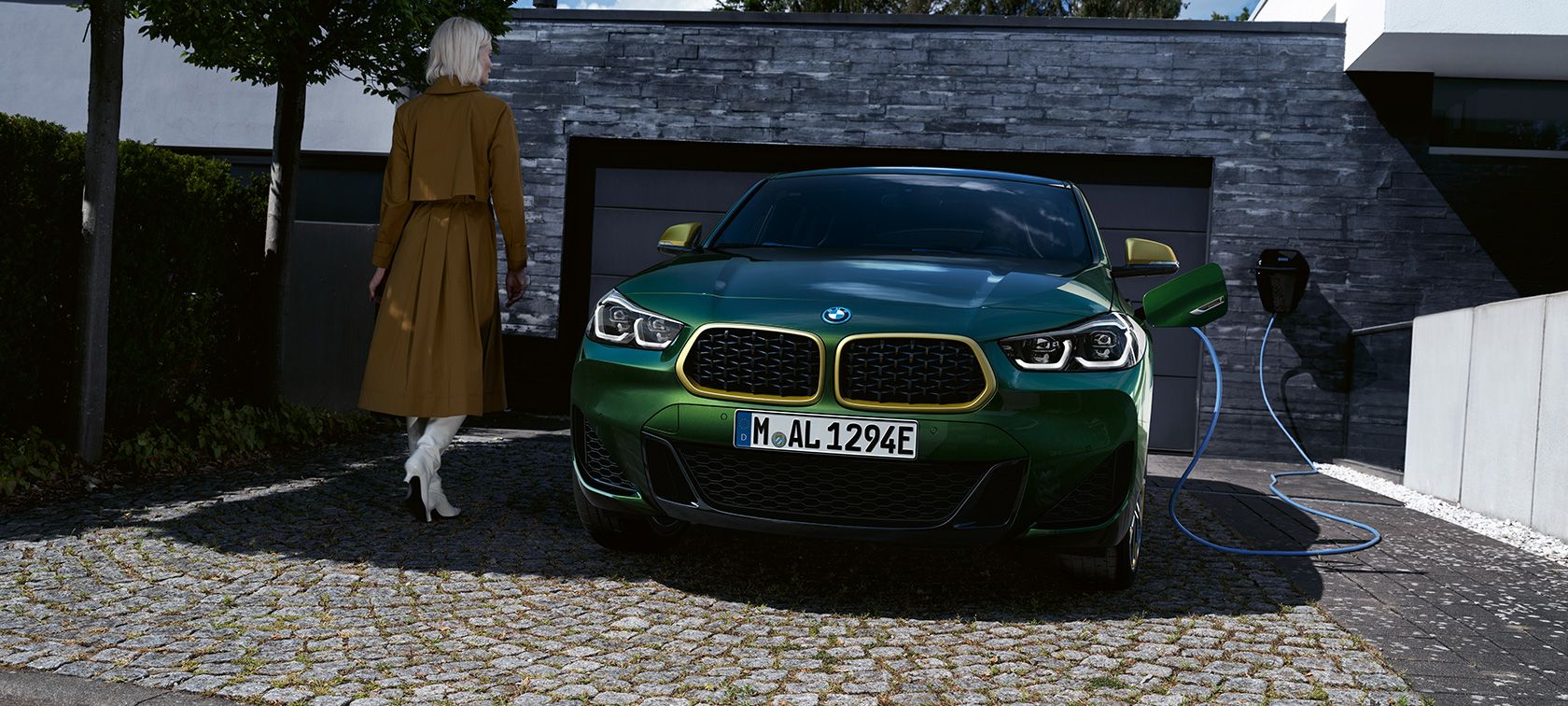 Grüner BMW X2 mit angeschlossenem Ladekabel vor einer Garage neben einer Frau Grüner BMW X2 mit angeschlossenem Ladekabel vor einer Garage neben einer Frau.