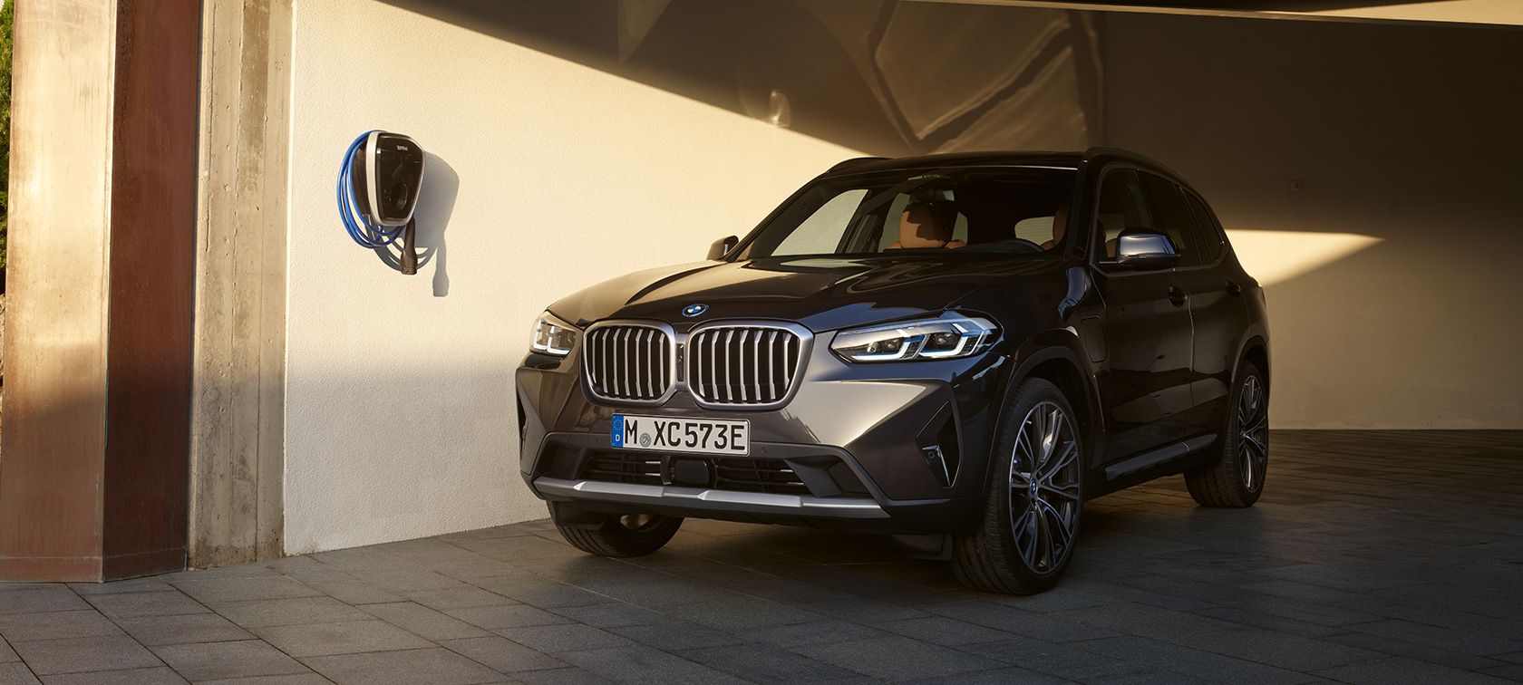 Dunkler BMW X3 mit angeschlossenem Ladekabel neben einer Wallbox Dunkler BMW X3 mit angeschlossenem Ladekabel neben einer Wallbox.