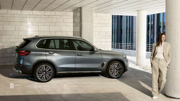Silberner BMW X5 in Seitenansicht neben einer Frau vor einem modernen Gebäude Silberner BMW X5 in Seitenansicht neben einer Frau vor einem modernen Gebäude.