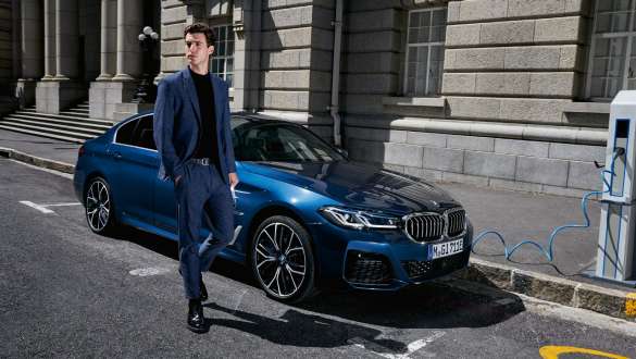 Mann geht neben einer blauen BMW 5er Limousine an Ein Mann geht neben einer blauen BMW 5er Limousine an einer öffentlichen Ladestation vorbei.