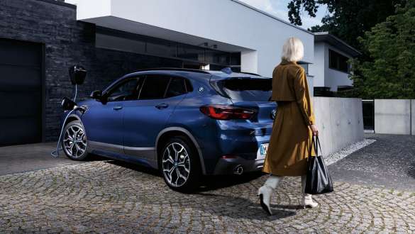 Frau geht neben einem blauen BMW X2 mit angeschlossenem Eine Frau geht neben einem blauen BMW X2 mit angeschlossenem Ladekabel vor einem Haus vorbei.