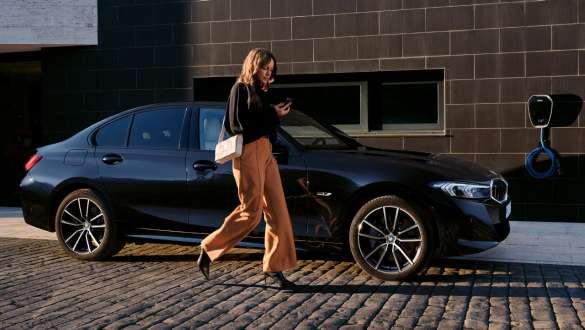 Frau geht neben einer dunklen BMW 3er Limousine mit Eine Frau geht neben einer dunklen BMW 3er Limousine mit angeschlossenem Ladekabel vorbei.