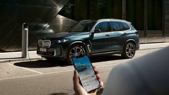 Hand hält ein Smartphone vor einem BMW X5 an einer Ladesäule Eine Hand hält ein Smartphone vor einem BMW X5 an einer Ladesäule.